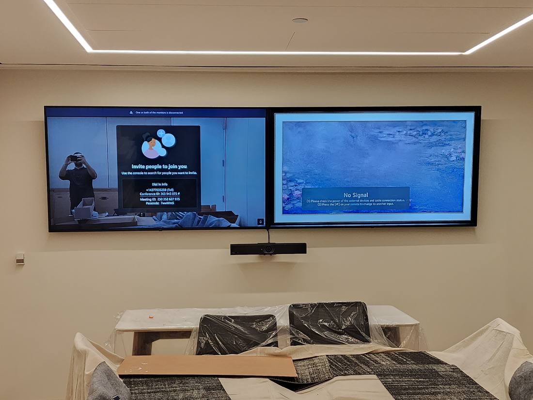 Two wall-mounted displays in a boardroom, one showing a conferencing invite screen and one showing no signal