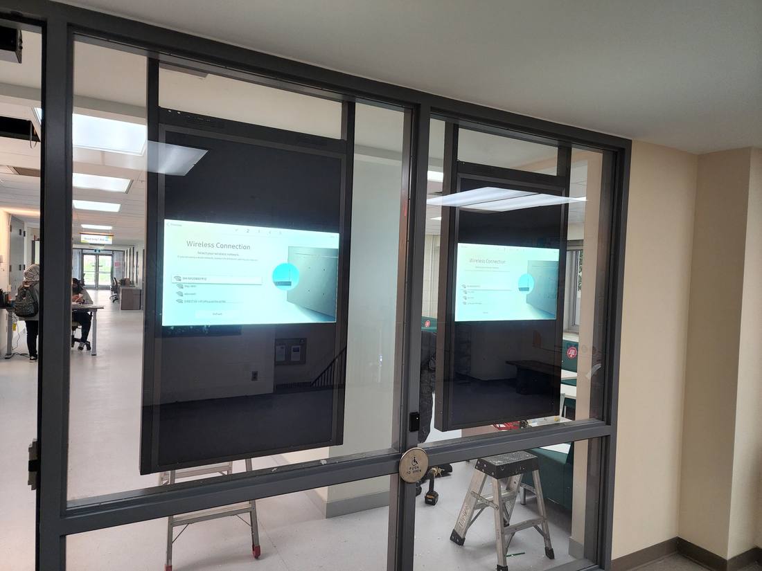 Two digital signage displays showing wireless connection setup screens behind a glass corridor wall